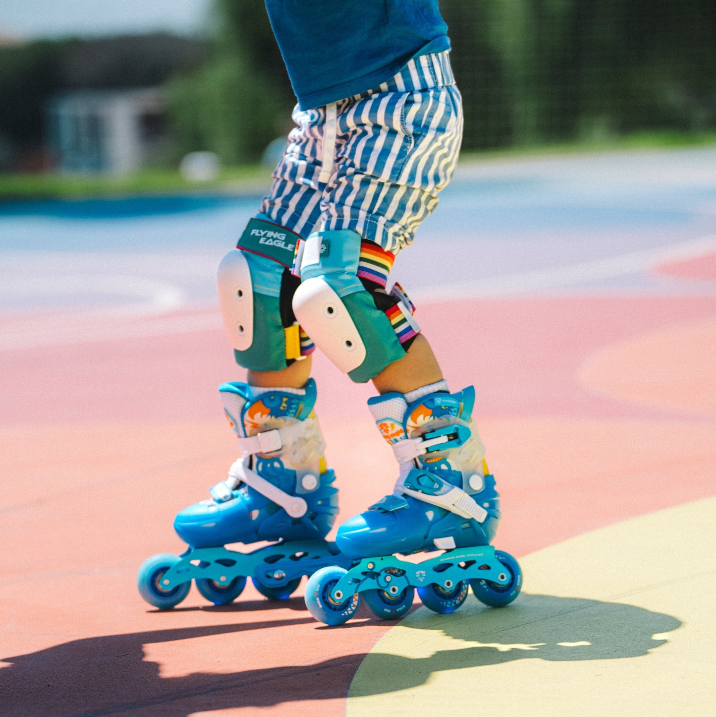 A close-up of a child wearing Flying Eagle L6 Lumos Kids Inline Skates with matching Vallant Protectors knee pads and striped shorts on a colorful outdoor court.