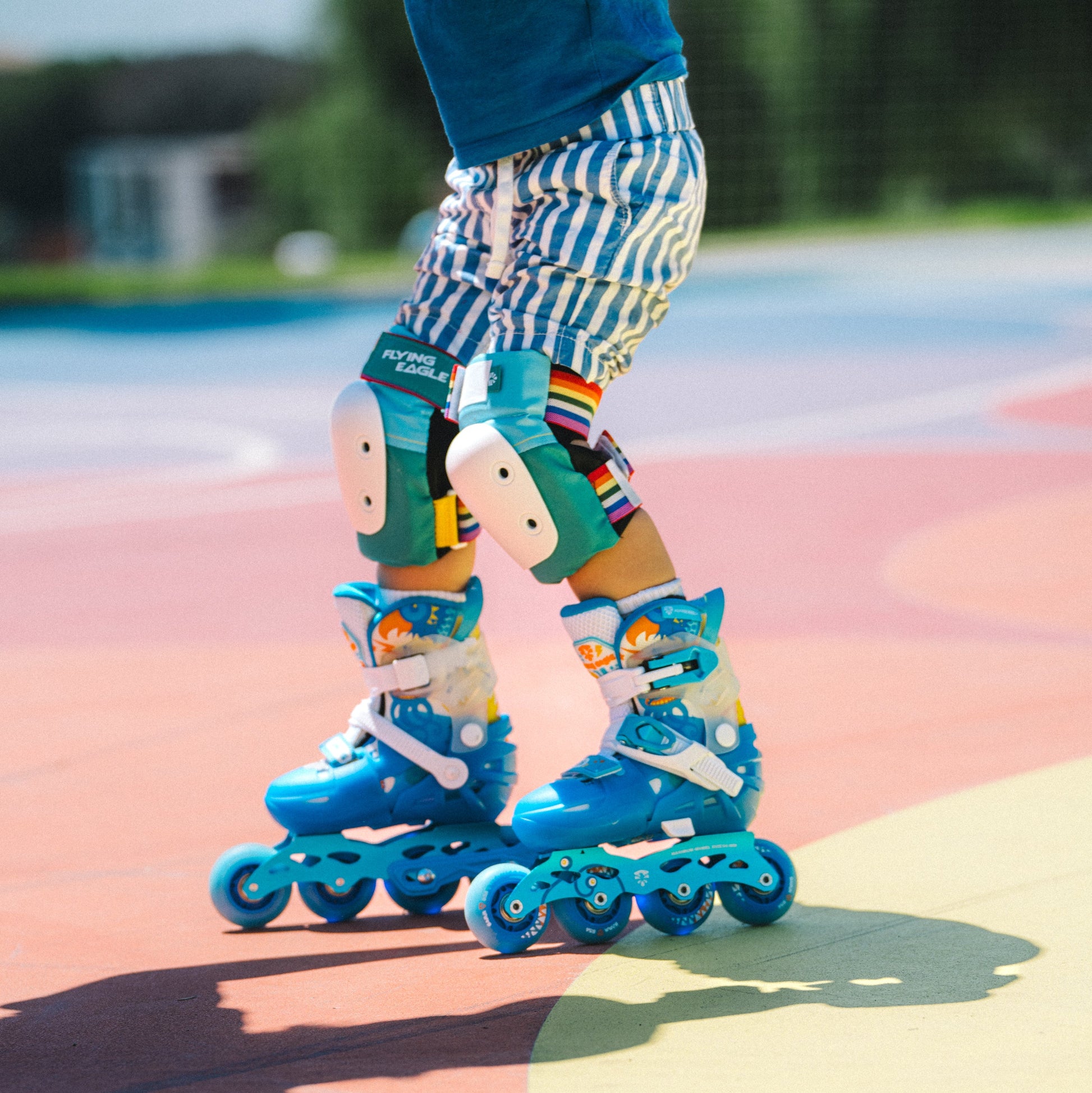 A close-up of a child wearing Flying Eagle L6 Lumos Kids Inline Skates with matching Vallant Protectors knee pads and striped shorts on a colorful outdoor court.