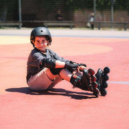 A child wearing RIDER helmet, protective pads, and Flying Eagle NIMBUS PRO black kids inline skates sits on a red outdoor court, looking toward the camera.
