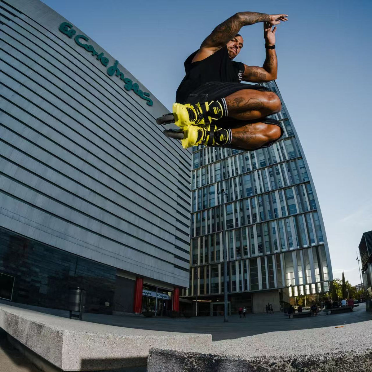 A man wearing a Flying Eagle REAVER S7680 Yellow Black adult inline skates 4 wheels 85A rockerable axles jumps in mid-air over a concrete ledge in an urban setting.