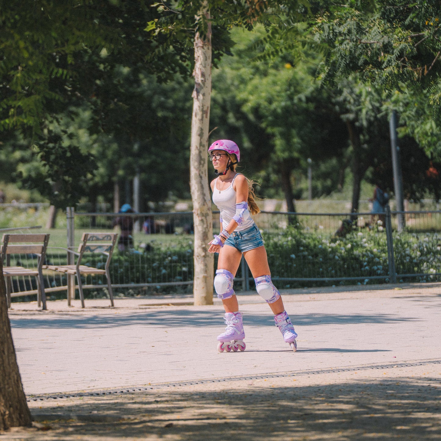 A young girl wearing Flying Eagle SHRIKE inline skates, including URBAN Pink Cloud helmet, knee pads, elbow pads, and pink roller skates, is skating in a sunny park.
