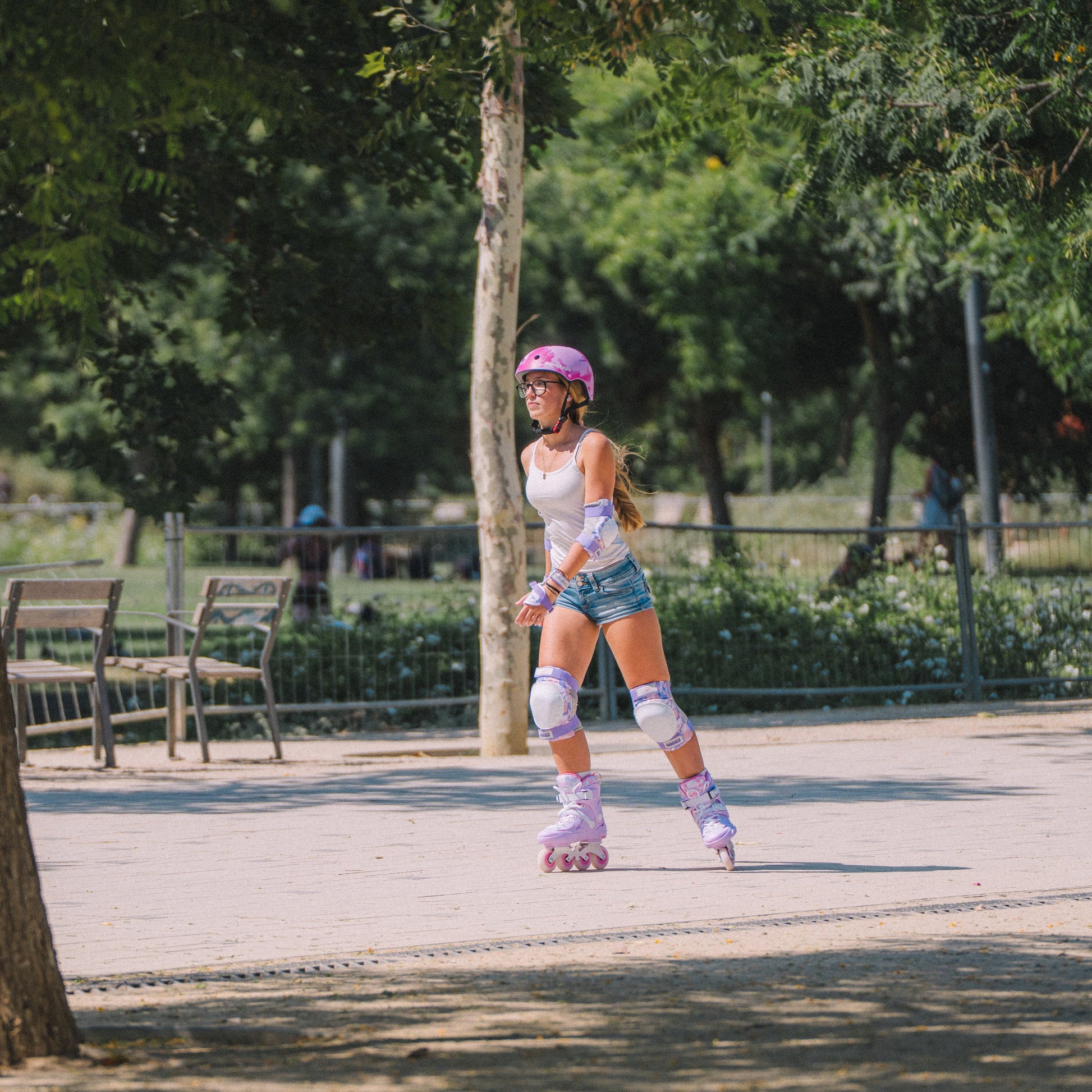 A young girl wearing Flying Eagle SHRIKE inline skates, including URBAN Pink Cloud helmet, knee pads, elbow pads, and pink roller skates, is skating in a sunny park.